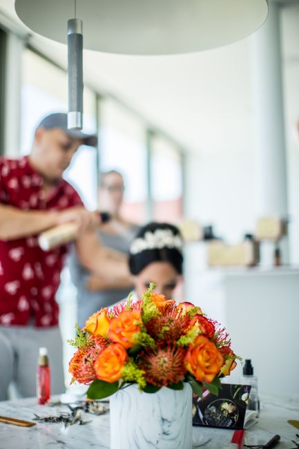 A vibrant floral arrangement featuring orange roses, red and pink proteas, and green foliage placed in a white marble-patterned vase. The bouquet is positioned on a white table in a bright, modern ind