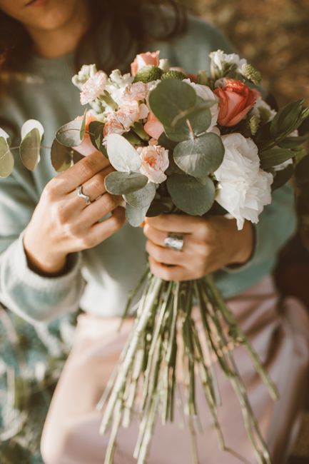 A person holding a delicate bouquet of fresh flowers with a mix of soft pink roses, white peonies, and eucalyptus leaves. The arrangement features a natural, loose style with visible green stems bound