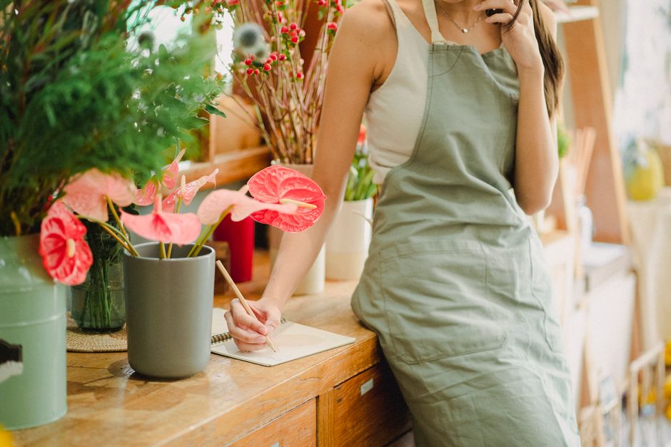 A florist in a beige apron is standing behind a wooden counter in a well-lit flower shop, taking notes on a notepad. On the counter, there are various floral arrangements, including a bouquet of vibra