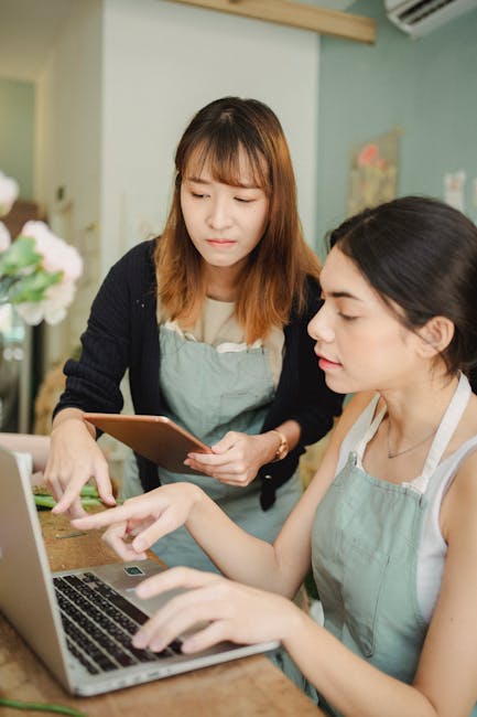 The image features two women in a floral shop, with one woman holding a tablet and the other working on a laptop. The woman standing has shoulder-length light brown hair with bangs, wearing a navy car