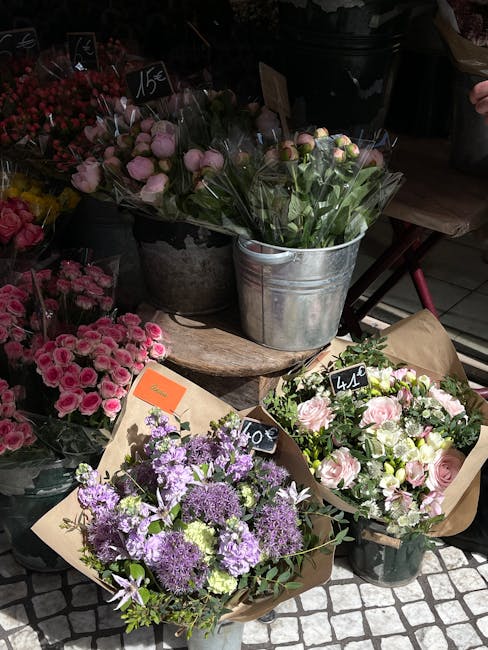 An arrangement of fresh flowers displayed outdoors at a flower shop, featuring pink roses and light pink peonies wrapped in brown paper, alongside purple and lavender blossoms in a bouquet. The flower